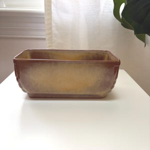 May include: A rectangular ceramic planter with a brown and yellow ombre glaze. The planter has a slightly textured surface and a raised rim. The planter is empty and sits on a white surface.