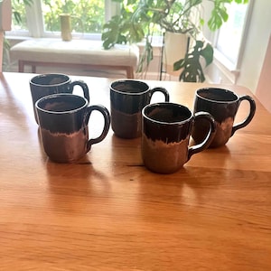 May include: Five ceramic mugs with a dark brown and black glaze are arranged on a wooden table. The mugs have a rounded shape with a handle on the side. The interior of the mugs is a dark brown color.