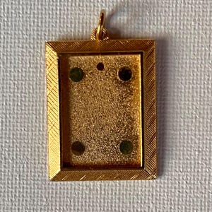 May include: Gold-toned rectangular pendant with a textured surface and a small loop for a chain. The pendant has a decorative border and four dark green and black dots. A small red dot is also visible.