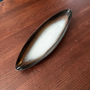 May include: A long, oval-shaped serving dish with a white glaze and brown speckles. The dish is made of ceramic and has a glossy finish.