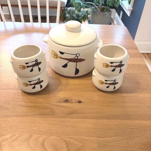 May include: A cream-colored ceramic serving set featuring a lidded casserole dish and two sets of stacked bowls. Each piece is adorned with a brown, black, and yellow fork and spoon design. The set is displayed on a wooden table.
