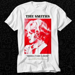 Puede incluir: Camiseta blanca con un gráfico rojo y blanco del perfil de una mujer, con el nombre de la banda "THE SMITHS" arriba. Debajo está el texto "SHEILA TAKE A BOW" y otro texto. La camiseta es de cuello redondo.