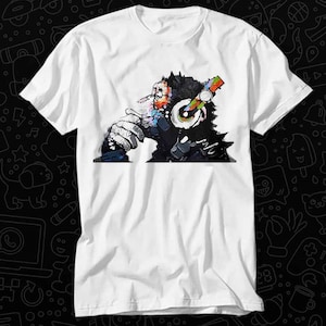 Peut inclure: T-shirt blanc avec un imprimé graphique coloré d'un singe portant un casque. Le singe regarde vers la droite et a une expression pensive.