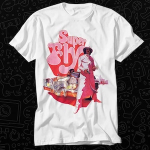 May include: White t-shirt featuring a graphic print of a man in a white suit and red coat standing next to a gold car. The text "Super Fly" is printed in pink and red letters.