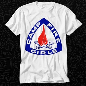 Peut inclure: T-shirt blanc avec un logo Camp Fire Girls bleu et rouge. Le logo est un triangle avec les mots "Camp Fire Girls" autour et une flamme rouge avec des bûches croisées à l'intérieur.