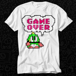 May include: White t-shirt with a pixelated green character and a speech bubble that says "GAME OVER" in pink.