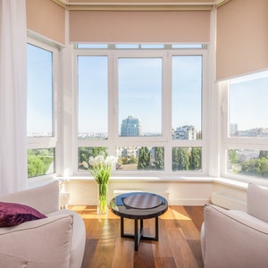 May include: Two white armchairs with a purple throw pillow each, facing a round wooden table with a black top. The chairs and table are in front of a large bay window with white blinds and a view of a city skyline.