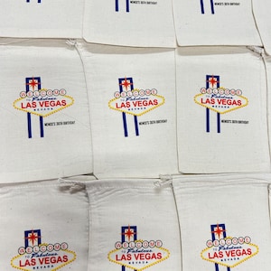 Puede incluir: Bolsas de tela blancas con un dise&ntilde;o impreso del ic&oacute;nico letrero "Welcome to Fabulous Las Vegas" en rojo, amarillo y azul. El texto "MEMOS'S 36TH BIRTHDAY" est&aacute; impreso debajo. Las bolsas est&aacute;n dispuestas en una cuadr&iacute;cula.
