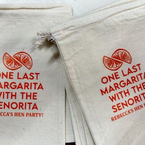 Puede incluir: Bolsas de lona color crema con texto rojo y un gr&aacute;fico de rodaja de c&iacute;trico. El texto dice "ONE LAST MARGARITA WITH THE SENORITA REBECCA'S HEN PARTY!" Ideal para una despedida de soltera.