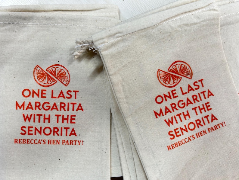 Puede incluir: Bolsas de lona color crema con texto rojo y un gr&aacute;fico de rodaja de c&iacute;trico. El texto dice "ONE LAST MARGARITA WITH THE SENORITA REBECCA'S HEN PARTY!" Ideal para una despedida de soltera.