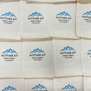 Puede incluir: Bolsas de tela blanquecina con un dise&ntilde;o de monta&ntilde;a azul y el texto "ALTITUDE KIT JARED & EMILY 10.26.2025". Las bolsas est&aacute;n dispuestas en una cuadr&iacute;cula, posiblemente para un evento especial.