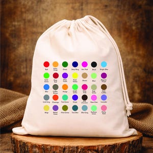 Puede incluir: Una bolsa con cord&oacute;n de color crema muestra una carta de colores con varios tonos. Cada color est&aacute; representado por un peque&ntilde;o c&iacute;rculo con su nombre debajo, incluyendo tonos de rojo, verde, azul y morado. La bolsa est&aacute; sobre una superficie de madera.