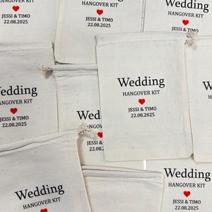 Puede incluir: Varios bolsos de tela blanquecina con cordones. Cada bolso tiene impreso en negro las palabras "Wedding HANGOVER KIT", un coraz&oacute;n rojo y los nombres "JESSI & TIMO" con la fecha "22.08.2025".