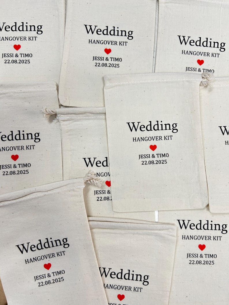 Puede incluir: Varios bolsos de tela blanquecina con cordones. Cada bolso tiene impreso en negro las palabras "Wedding HANGOVER KIT", un coraz&oacute;n rojo y los nombres "JESSI & TIMO" con la fecha "22.08.2025".