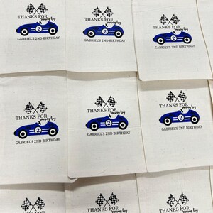 Puede incluir: Bolsas de tela color crema para recuerdos de fiesta con un gr&aacute;fico de coche de carreras azul, banderas a cuadros y el texto "THANKS FOR racing by" y "GABRIEL'S 2ND BIRTHDAY". Las bolsas est&aacute;n dispuestas en forma de cuadr&iacute;cula.