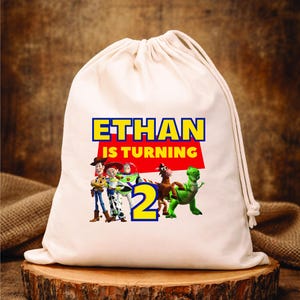 Puede incluir: Una bolsa con cordón de color crema con el texto "ETHAN IS TURNING 2" en azul y amarillo. La bolsa está decorada con imágenes de personajes de la película de animación Toy Story. La bolsa está sobre una superficie de madera.