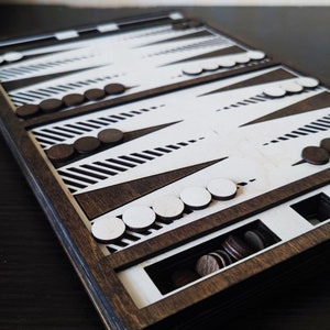 May include: A wooden backgammon game set with black and white playing pieces. The board is made of dark wood with white stripes and black triangles. The pieces are stored in a compartment on the side of the board.