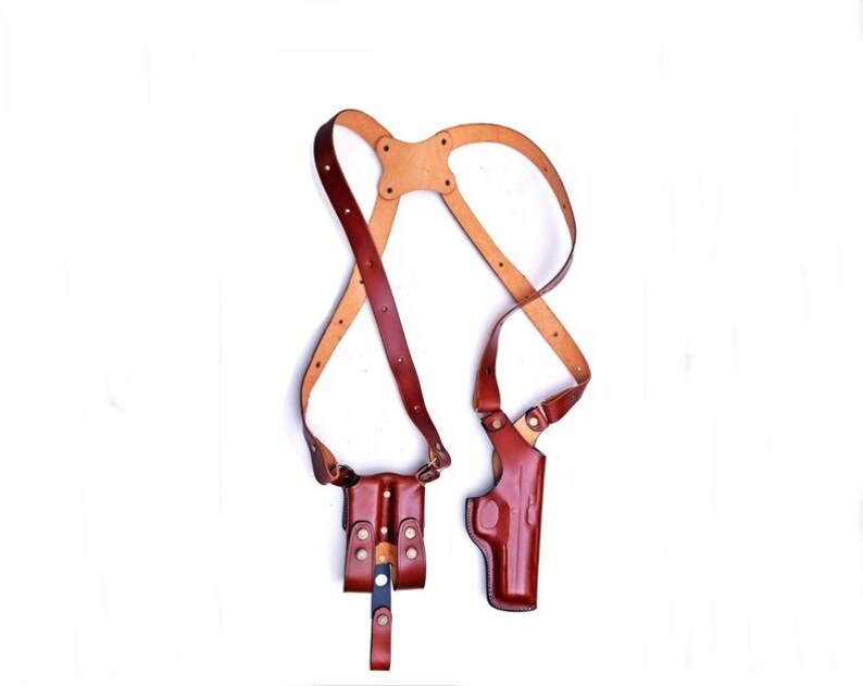 May include: A brown leather shoulder holster with two holsters, one for a handgun and one for a magazine. The holster has adjustable straps and is designed to be worn across the body.