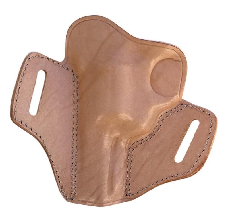 May include: A light brown leather holster with a molded shape. The holster features two slots for a belt and is stitched with dark thread around the edges. The leather has a natural, unpainted finish.