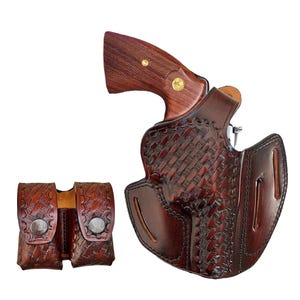 Può includere: Una fondina in pelle marrone con un motivo intrecciato, che contiene un revolver con un'impugnatura in legno. È presente anche una doppia tasca per caricatori in pelle marrone abbinata. Gli oggetti sono su sfondo bianco.