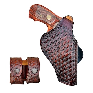 Op de afbeelding: Een bruin leren holster met een geweven patroon, met een revolver met een houten handvat. Een bijpassend bruin leren etui met twee compartimenten en zilveren drukknopen is ook zichtbaar.