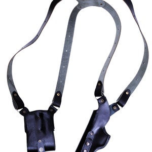 May include: A black leather holster with a shoulder strap. The holster has two compartments, one for a gun and one for a magazine. The strap is adjustable and has a buckle.