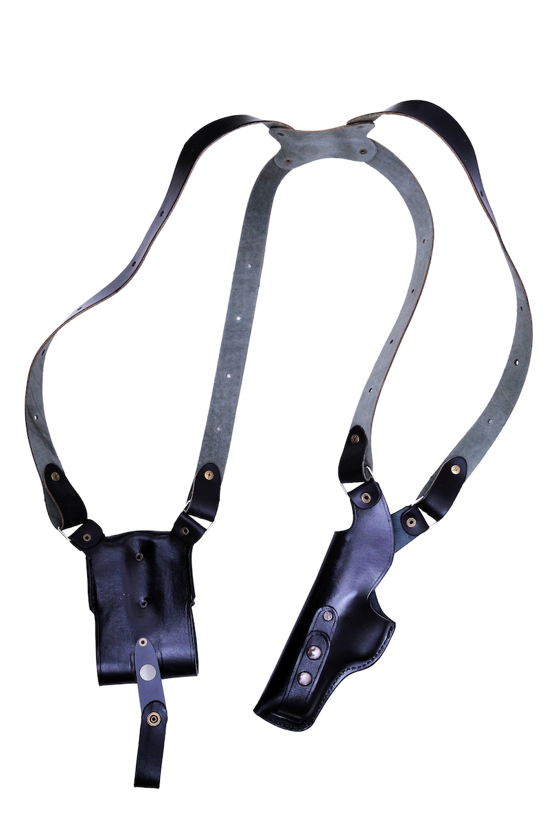 May include: A black leather holster with a shoulder strap. The holster has two compartments, one for a gun and one for a magazine. The strap is adjustable and has a buckle.