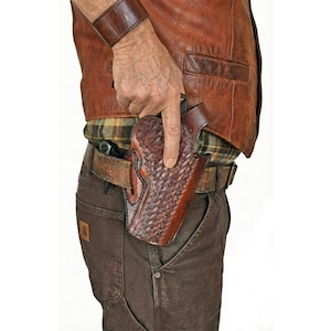 May include: A brown leather holster with a woven pattern, worn on a brown leather belt. The holster is attached to a brown leather vest and brown pants. A brown leather wristband is also visible.