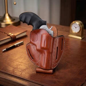 Op de afbeelding: Een bruin lederen holster met een zilverkleurige revolver met een zwarte, getextureerde handgreep. Het holster is voorzien van zichtbare witte stiksels langs de randen en meerdere riemsleuven om het vuurwapen stevig vast te houden.