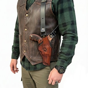 May include: Brown leather shoulder holster with a matching brown leather gun. The holster has a paw print design. The person is wearing a brown leather vest and a green plaid shirt.