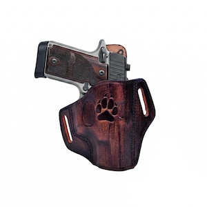 Puede incluir: Una funda de cuero marrón con un diseño de huella de pata, que sostiene una pistola plateada y negra. La funda tiene un acabado oscuro y desgastado y costuras visibles. El arma de fuego es parcialmente visible.