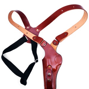 K&ouml;nnte beinhalten: Ein braunes Leder-Schulterholster mit einem schwarzen Riemen. Das Holster hat eine Klappe, die die Waffe bedeckt, und einen Druckknopfverschluss.