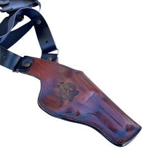 Puede incluir: Una funda de hombro de cuero marr&oacute;n con un dise&ntilde;o de huella de pata. La funda tiene un acabado marr&oacute;n oscuro con correas negras y herrajes met&aacute;licos. El cuero es texturizado y parece hecho a mano.