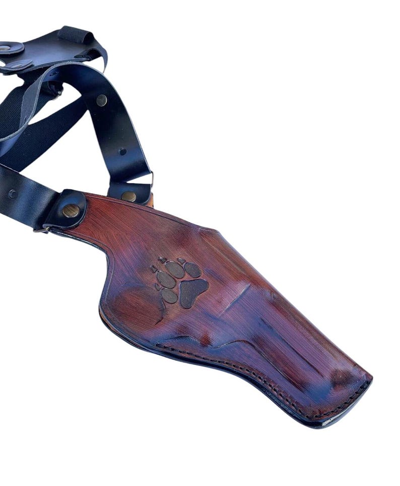 Puede incluir: Una funda de hombro de cuero marr&oacute;n con un dise&ntilde;o de huella de pata. La funda tiene un acabado marr&oacute;n oscuro con correas negras y herrajes met&aacute;licos. El cuero es texturizado y parece hecho a mano.