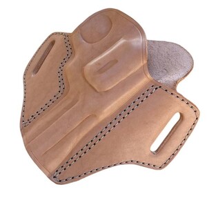 May include: A light brown leather holster with black stitching. The holster has a molded shape with a curved design and two slots for a belt. The edges are reinforced with black stitching.