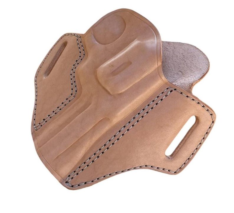 May include: A light brown leather holster with black stitching. The holster has a molded shape with a curved design and two slots for a belt. The edges are reinforced with black stitching.
