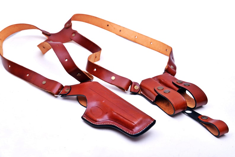 May include: A brown leather shoulder holster with two separate compartments for carrying a firearm and extra ammunition. The holster has adjustable straps and a secure closure.
