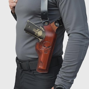 May include: A brown leather shoulder holster with a skull design, holding a black handgun with a skull emblem. The holster is worn over a gray long-sleeve shirt and black pants.