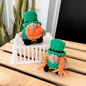 May include: Two crocheted leprechaun figures with green hats, orange beards, and black belts. The figures are sitting on a white basket and a wooden surface.