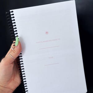 May include: A white spiral bound planner with a pink flower design and the text "This planner belongs to" and "Year" printed on the cover.