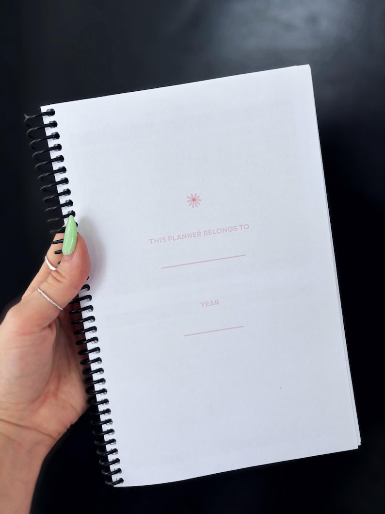 May include: A white spiral bound planner with a pink flower design and the text "This planner belongs to" and "Year" printed on the cover.