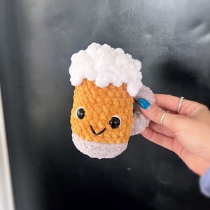 Op de afbeelding: Een handgemaakte, gehaakte bierpul met een lachend gezicht. De pul is voornamelijk oranje met een witte, donzige bovenkant die schuim voorstelt. De onderkant van de pul is grijs en heeft een klein grijs handvat. De pul wordt tegen een donkere achtergrond gehouden.