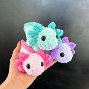 Op de afbeelding: Drie gehaakte axolotl knuffels in roze, blauw en paars. De knuffels hebben grote ogen en zijn gemaakt van zacht, pluche garen.