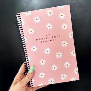 May include: A pink spiral bound notebook with a white daisy pattern and the text "MY MARKET PREP PLANNER" in brown lettering.