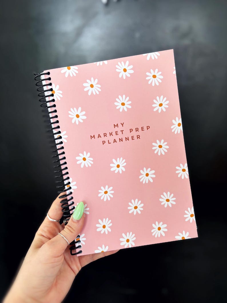 May include: A pink spiral bound notebook with a white daisy pattern and the text "MY MARKET PREP PLANNER" in brown lettering.