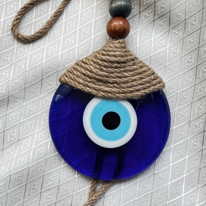 Peut inclure: Un charme bleu de mauvais œil avec un œil blanc et bleu clair, entouré d'une corde brune, suspendu à une corde brune avec des perles en bois.