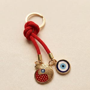 May include: A gold-coloured keyring with a red cord and a gold ring. The keyring features a pomegranate charm and an evil eye charm, both with blue and white details. The pomegranate charm has a red base.