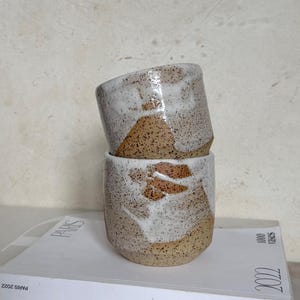 May include: Two handmade ceramic cups stacked. The cups have a speckled tan base with white and brown glaze details. The cups are on a white book with "PARIS" printed on the cover. The book is from 2022.