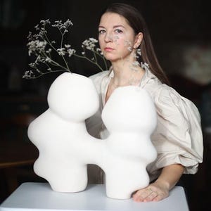 Könnte beinhalten: Eine weiße Keramikvase mit einem abstrakten, miteinander verbundenen Design. Die Vase enthält zarte weiße Blumen. Das Bild ist vor einem dunklen Hintergrund platziert, mit einer Person im Hintergrund.