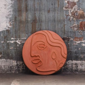 Könnte beinhalten: Runde Wandkunst aus Terrakotta mit einem stilisierten Gesichtsprofil. Es lehnt an einer verwitterten Wand mit abblätternder blauer, weißer und brauner Farbe. Das Kunstwerk ist ein dekoratives Element, geeignet für die Inneneinrichtung.